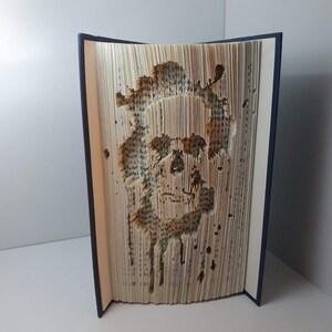 May include: A book sculpture featuring a skull design cut into the pages. The skull is brown with a dripping effect. The book has a dark blue spine and is displayed upright on a white surface.