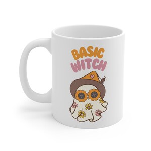 May include: White ceramic mug featuring a ghost wearing a witch's hat and sunglasses. The text "BASIC WITCH" is printed on the mug.
