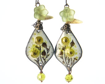 Può includere: Un paio di orecchini pendenti con una forma a goccia di smalto bianco con un motivo floreale con fiori gialli e steli marroni. Gli orecchini hanno una perla verde a forma di stella e una perla di cristallo verde nella parte inferiore.