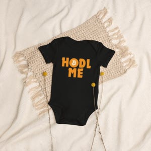 May include: Black baby onesie with the text "HODL ME" in orange letters. A yellow Bitcoin symbol is in the middle of the word "HODL".