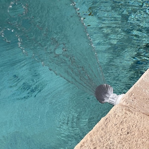 Puede incluir: Primer plano de una boquilla de fuente de piscina blanca que rocía agua en una piscina turquesa.