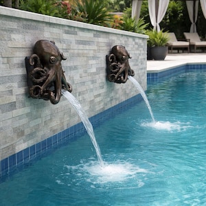 May include: Two bronze-colored octopus wall fountains mounted on a light gray stone wall, each featuring a detailed octopus sculpture with tentacles extending outward, releasing a steady stream of water into a blue swimming pool below.