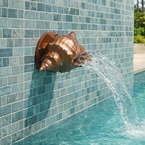 May include: A metallic copper-toned conch shell fountain spout mounted on a light blue tiled wall, discharging a steady stream of clear water into a swimming pool. The shell features a spiral design and a circular base plate attached to the wall.