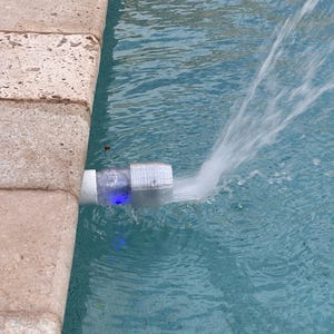 May include: A white cylindrical pool fountain attachment with a translucent section emitting a blue light, connected to a pipe at the edge of a swimming pool, projecting a steady stream of water into the pool basin with visible ripples.