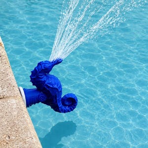 Könnte beinhalten: Ein leuchtend blauer, seepferdchenförmiger Pool-Fontänenaufsatz, der am Rand eines Schwimmbeckens befestigt ist. Das Seepferdchen ist horizontal ausgerichtet, wobei Wasser aus seinem Maul in den Pool sprüht und einen kontinuierlichen Wasserstrahl erzeugt.