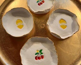 May include: Four white ceramic trinket dishes with gold trim. Each dish features a different painted design: a lemon, a cherry, a lemon, and a cherry.