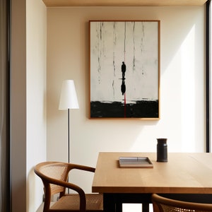 May include: A minimalist dining room with a wooden table and chairs. A framed black and white abstract painting hangs on the wall. A lamp with a white shade stands in the corner.