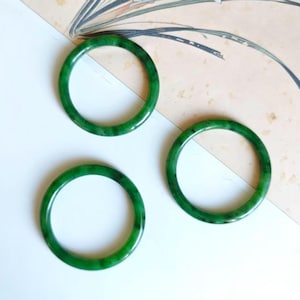 May include: Three polished, jade green bangles. The circular bracelets are a vibrant, translucent green, showcasing natural variations in colour. The bangles are displayed on a white and cream-coloured surface.