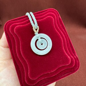 May include: A pale, translucent circular jade pendant featuring a smaller concentric ring in the center. The pendant is suspended from a light gray braided cord, which is secured at the top by a small, metallic gold-toned wire wrapping detail.