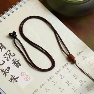 May include: A dark brown necklace with a sliding knot and wooden bead accents. The necklace is lying on a calendar with Chinese characters. The calendar has dates and days of the week in Chinese.