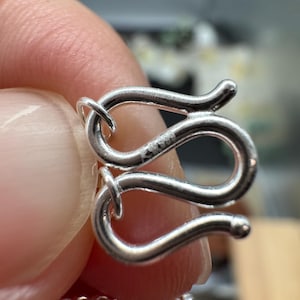 Puede incluir: Un cierre de joyer&iacute;a en forma de S de plata con un acabado met&aacute;lico pulido. El cierre presenta un dise&ntilde;o curvo y serpentino con un peque&ntilde;o sello grabado S999 visible en la superficie. El gancho est&aacute; conectado a un eslab&oacute;n de cadena fino.