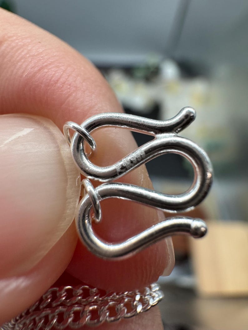 Puede incluir: Un cierre de joyer&iacute;a en forma de S de plata con un acabado met&aacute;lico pulido. El cierre presenta un dise&ntilde;o curvo y serpentino con un peque&ntilde;o sello grabado S999 visible en la superficie. El gancho est&aacute; conectado a un eslab&oacute;n de cadena fino.