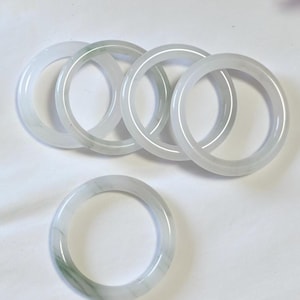 May include: Six jadeite jade bangles. The bangles are a translucent white with subtle green veining. The circular bracelets are displayed on a white surface. The jadeite bangles are a classic jewellery item.