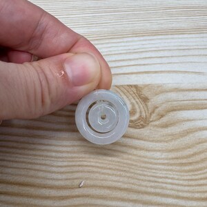 May include: A pale, translucent white jade pendant featuring a circular outer ring and a smaller, concentric circular inner ring connected by a thin, clear filament. The smooth, polished stone has a subtle, cloudy appearance with soft grey tones.