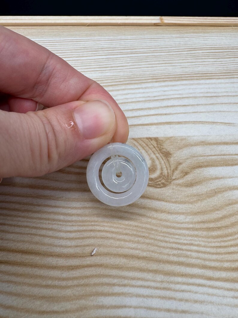 May include: A pale, translucent white jade pendant featuring a circular outer ring and a smaller, concentric circular inner ring connected by a thin, clear filament. The smooth, polished stone has a subtle, cloudy appearance with soft grey tones.