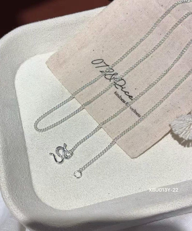 Puede incluir: Un collar de cadena en tono plateado con un dise&ntilde;o de eslabones delicados y un cierre de mosquet&oacute;n seguro. El collar se muestra sobre una bolsa de tela beige claro con el texto 07&Rice kindness for wellness y el c&oacute;digo XBU013Y-22.