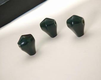 May include: Three dark green, faceted knobs with a glossy finish are arranged on a white surface. The knobs have a conical shape with a small hole at the top. The background includes a black object.