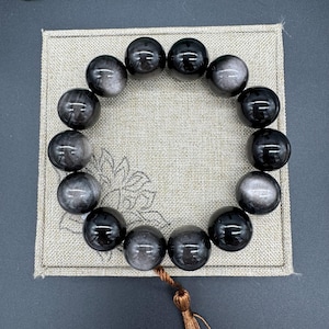 May include: A black obsidian bracelet with a brown tassel. The bracelet is made of round beads with a shimmering effect.