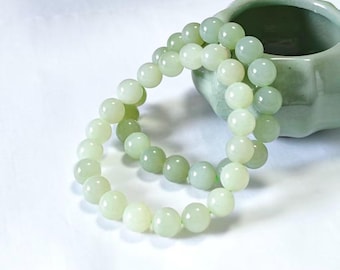 May include: Two light green jade bead bracelets. The bracelets are made of round, polished beads. A small, light green ceramic container is in the background. The bracelets are on a white surface.