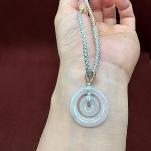 May include: A pendant featuring two concentric circular light-colored translucent stone rings suspended from a braided silver-toned cord. The cord includes a section of metallic gold-toned thread wrapping and a small gold-toned bead near the top.