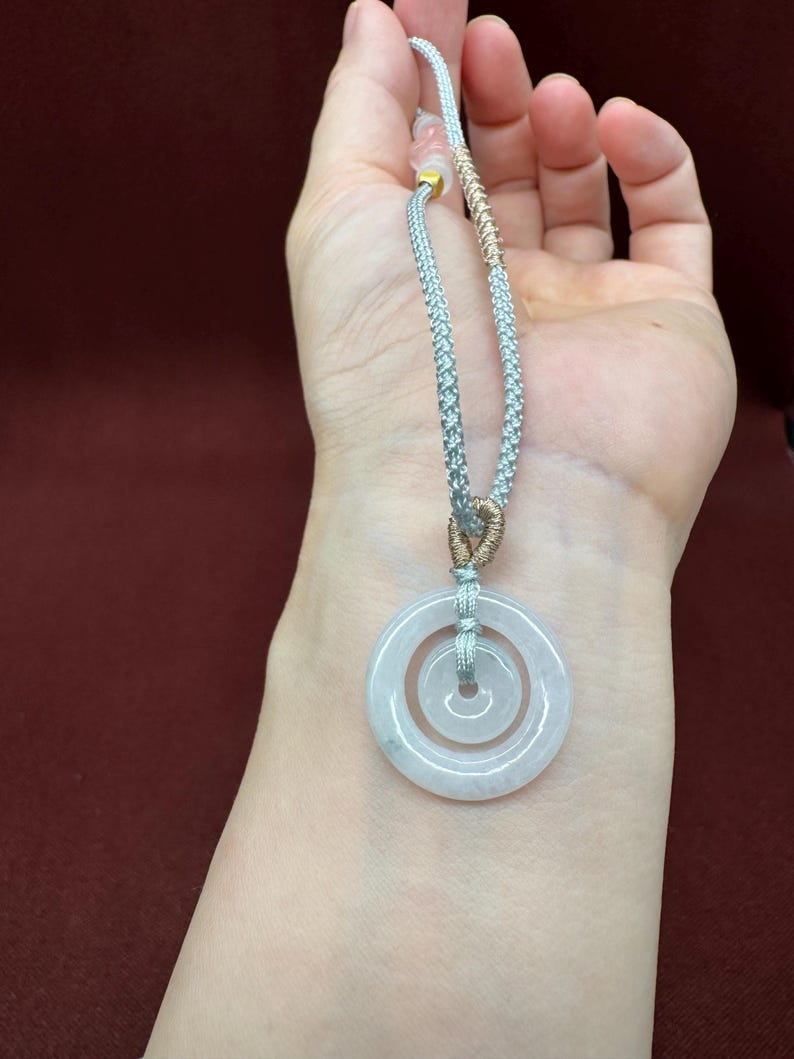 May include: A pendant featuring two concentric circular light-colored translucent stone rings suspended from a braided silver-toned cord. The cord includes a section of metallic gold-toned thread wrapping and a small gold-toned bead near the top.