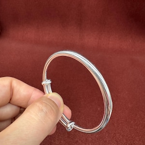 Może przedstawiać: Polerowana bransoletka typu bangle w kolorze srebrnym z gładką, zaokrągloną metalową obręczą. Konstrukcja zawiera regulowany mechanizm z dwoma małymi, cylindrycznymi elementami przesuwnymi, które pozwalają na zwiększenie lub zmniejszenie średnicy bransoletki.
