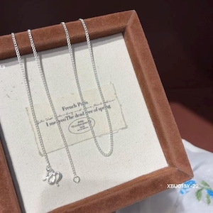 Puede incluir: Un collar de cadena en tono plateado con un dise&ntilde;o de eslabones delicados y un cierre decorativo. El collar se exhibe sobre un inserto de tela clara dentro de un marco marr&oacute;n, que incluye el texto French Paris y The dead tree of spring.