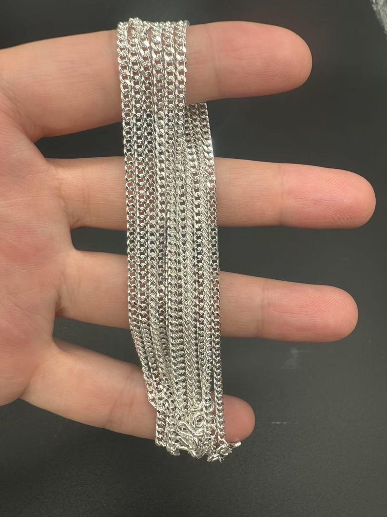 Puede incluir: Un collar de cadena de color plateado que presenta una serie de eslabones planos e interconectados que crean una textura met&aacute;lica continua y reflectante. La cadena est&aacute; compuesta por m&uacute;ltiples hilos con el mismo dise&ntilde;o de eslabones.