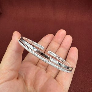 Pode incluir: Uma pulseira aberta de tom prateado com um design de banda dupla. Uma banda tem uma superfície lisa, polida e reflexiva, enquanto a banda adjacente apresenta um acabamento texturizado e cintilante. A pulseira é rígida.