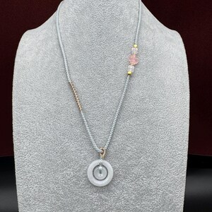 May include: A necklace featuring a light gray braided cord with a circular white stone pendant. The cord includes a rose gold-toned wire wrap, small gold-toned beads, and a translucent pink flower-shaped bead accent near the top right side.