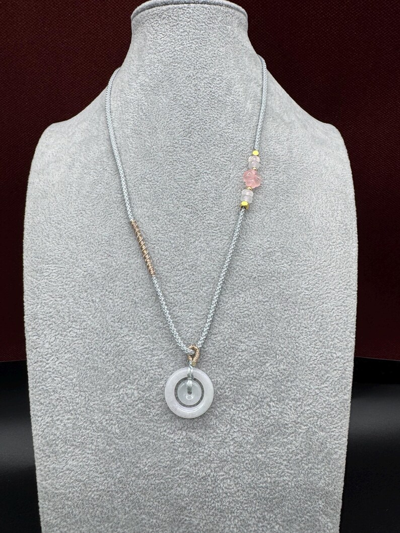 May include: A necklace featuring a light gray braided cord with a circular white stone pendant. The cord includes a rose gold-toned wire wrap, small gold-toned beads, and a translucent pink flower-shaped bead accent near the top right side.