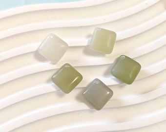 May include: Five square-shaped beads in varying shades of green and off-white. Each bead has a small hole at the top. The beads are arranged on a white, wavy surface.