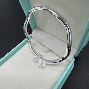 Pode incluir: Uma pulseira de bracelete prateada com um design ondulado. A pulseira apresenta um pequeno pingente de borboleta. A pulseira é exibida dentro de uma caixa de joias azul clara com interior branco. Um acessório elegante.