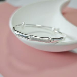 Pode incluir: Um bracelete de tom prateado com um design segmentado que imita a aparência de caules de bambu. O bracelete tem um acabamento polido e reflexivo com seções alternadas que exibem um detalhe de superfície sutilmente fosco e texturizado.