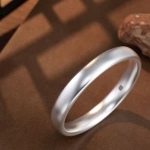 May include: A silver ring with a matte finish. The ring is smooth and rounded, with a simple, elegant design. The ring is set against a brown background with a piece of wood.