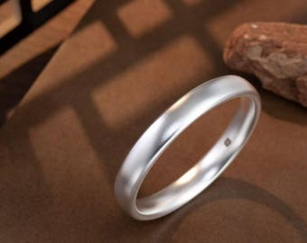 Puede incluir: Un anillo plateado con acabado mate. El anillo es liso y redondeado, con un diseño sencillo y elegante. El anillo está sobre un fondo marrón con un trozo de madera.