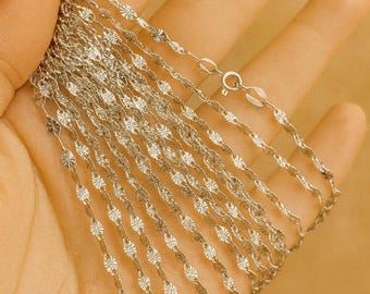Puede incluir: Se muestran varios collares delicados de plata con un diseño floral. Las cadenas se sostienen en una mano sobre un fondo cálido y neutro. Cada cadena presenta pequeños eslabones repetidos en forma de flor y se ve un cierre.