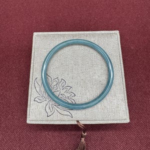May include: A teal-colored jade bangle bracelet rests on a square, light gray fabric box with a floral design. The box has a brown tassel attached. The background is a solid maroon color.