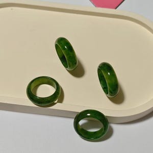 May include: Four green jade rings of varying sizes are displayed on a cream-colored tray. The rings have a smooth, polished surface and a translucent appearance, showcasing the natural variations in the jade's color.