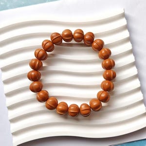 Puede incluir: Un brazalete de madera marrón con cuentas redondas. Las cuentas tienen un diseño tallado.