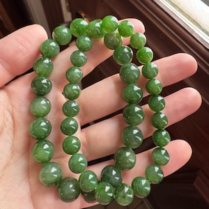 May include: Two strands of green jade bead bracelets. The beads vary in size and have a polished, glossy finish. The bracelets are held in a hand, showcasing the vibrant green color and the natural variations in the stone.