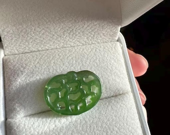 May include: A translucent green jade pendant in a heart shape with a carved pattern. The pendant is displayed inside a gray jewelry box with a soft, textured interior. The jade has a polished surface and is a vibrant shade of green.