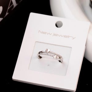 Peut inclure: Bague argentée ornée de deux motifs en forme de cœur et de petites pierres scintillantes. La bague est présentée sur une carte blanche avec l'inscription "New Jewelry". Il s'agit d'un bijou.