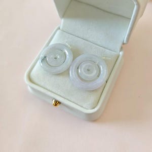 May include: Two circular, pale green jade pendants with a smooth, polished finish and a central hole, resting inside a white, rectangular jewelry box with a soft, light-colored interior lining and a small gold-toned clasp on the front edge.