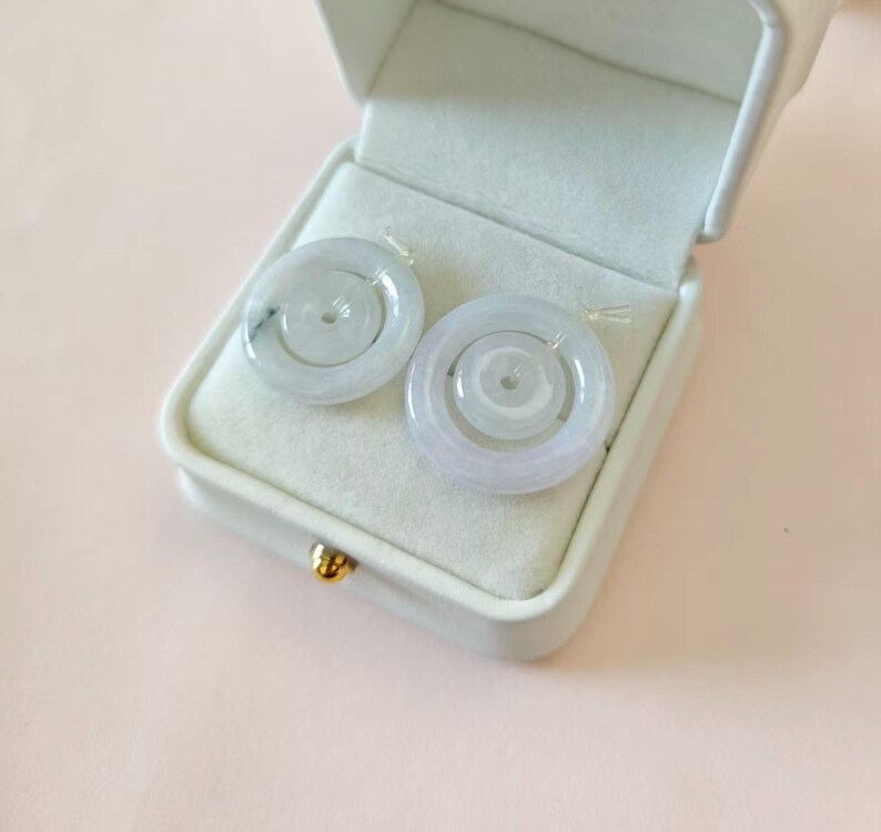 May include: Two circular, pale green jade pendants with a smooth, polished finish and a central hole, resting inside a white, rectangular jewelry box with a soft, light-colored interior lining and a small gold-toned clasp on the front edge.