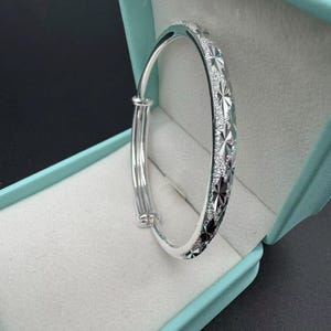 Pode incluir: Uma pulseira rígida de tom prateado com uma superfície texturizada e gravações repetitivas em forma de estrela. A pulseira possui um mecanismo deslizante ajustável e é apresentada dentro de uma caixa de joias azul-clara com forro macio.