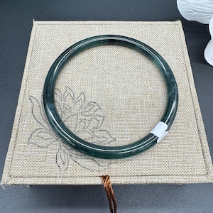 May include: A green jade bangle bracelet on a beige fabric background. The bangle is smooth and polished.