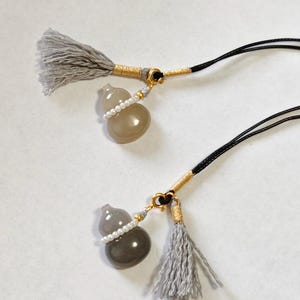 Op de afbeelding: Twee kalebasvormige hangers met grijze kwastjes en zwarte koorden. Elke hanger heeft een grijze, parelmoerachtige body, geaccentueerd met een rij kleine witte kralen. De koorden zijn bevestigd met goudkleurige metalen beslagen.
