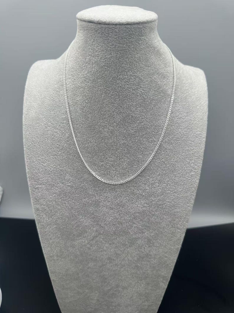 Puede incluir: Un collar de cadena de metal de color plateado con un dise&ntilde;o delicado de eslabones tipo barbada. El collar se muestra en una ca&iacute;da circular simple, exhibiendo los eslabones uniformes y reflectantes que forman un bucle met&aacute;lico continuo y pulido.