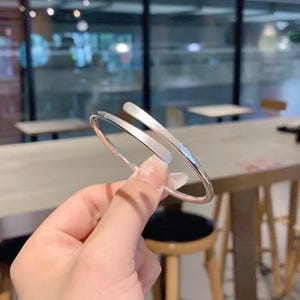 Pode incluir: Uma pulseira prateada com um design aberto. A pulseira tem um acabamento liso e polido e uma estética minimalista. As extremidades da pulseira se sobrepõem, criando um visual moderno e elegante. Esta joia é adequada para uso diário.